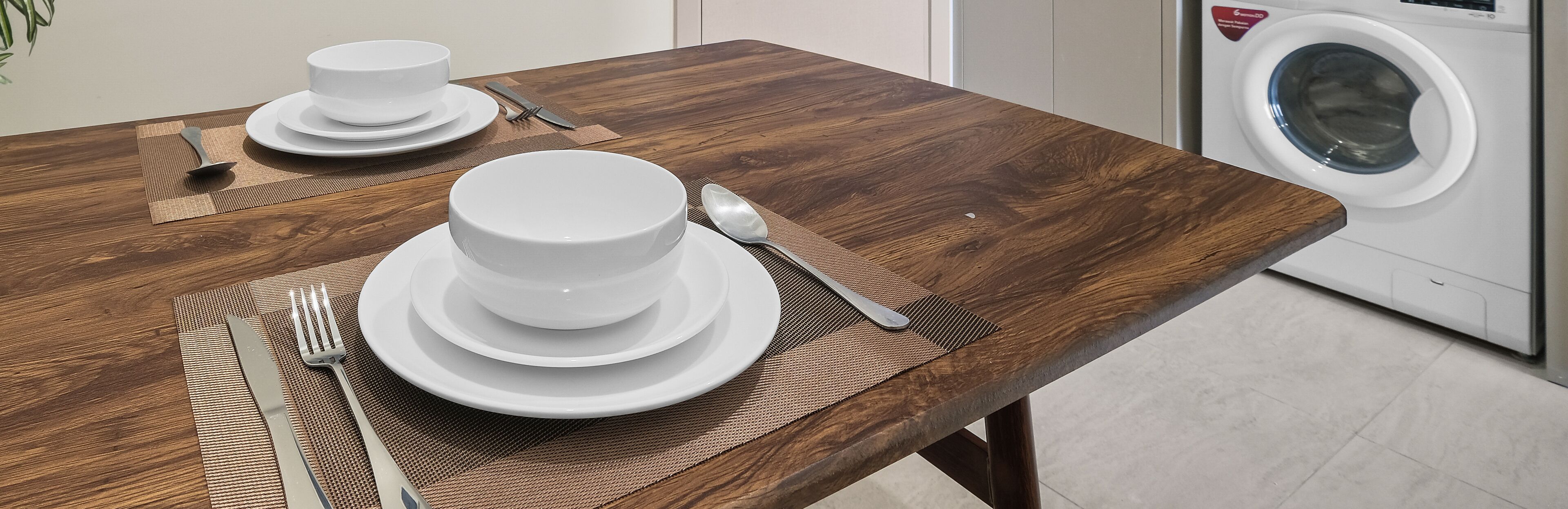 A dining table set with white plates, bowls, and cutlery, with a washing machine in the background.