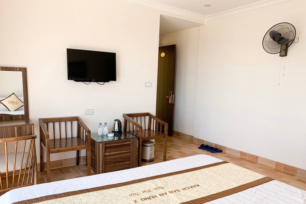 Habitación de hotel con cama, televisión, escritorio con sillas, hervidor de agua y ventilador.