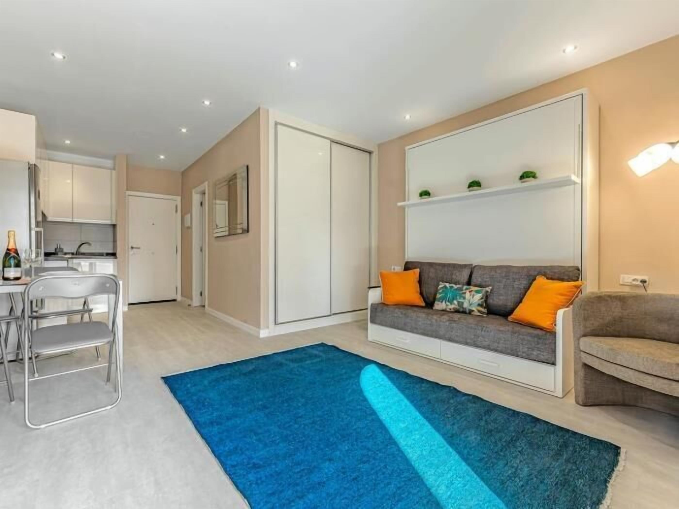 Ein modernes Wohnzimmer mit grauem Sofa, orangefarbenen Kissen, einem blauen Teppich und einer Küchenzeile mit weißen Schränken.