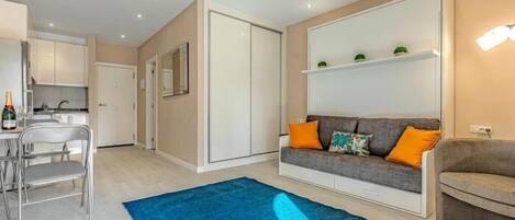 Ein modernes Wohnzimmer mit grauem Sofa, orangefarbenen Kissen, einem blauen Teppich und einer Küchenzeile mit weißen Schränken.