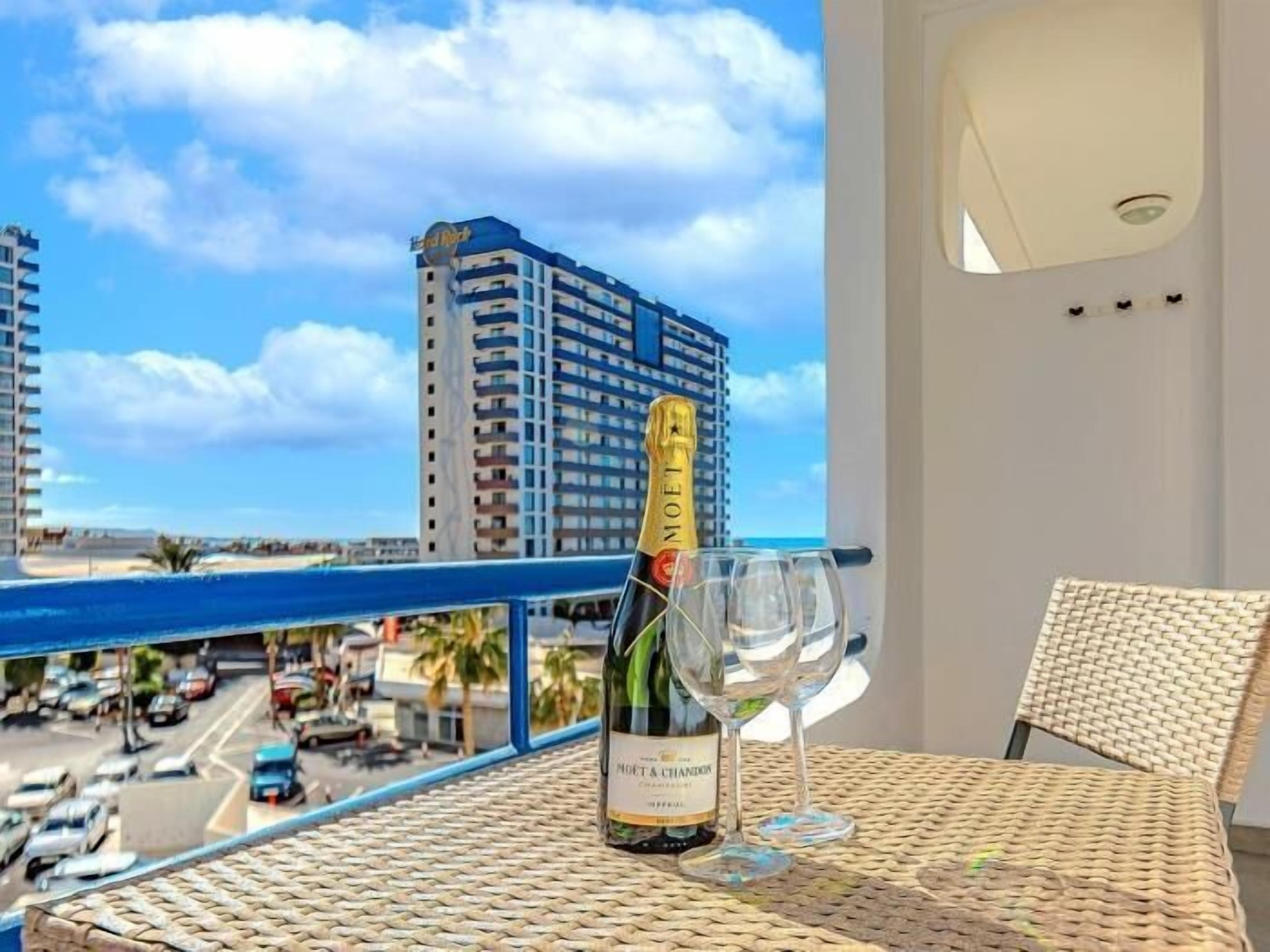 Uma varanda com uma garrafa de champanhe Moët & Chandon e duas taças, com vista para um horizonte urbano que inclui um prédio alto e uma praia.