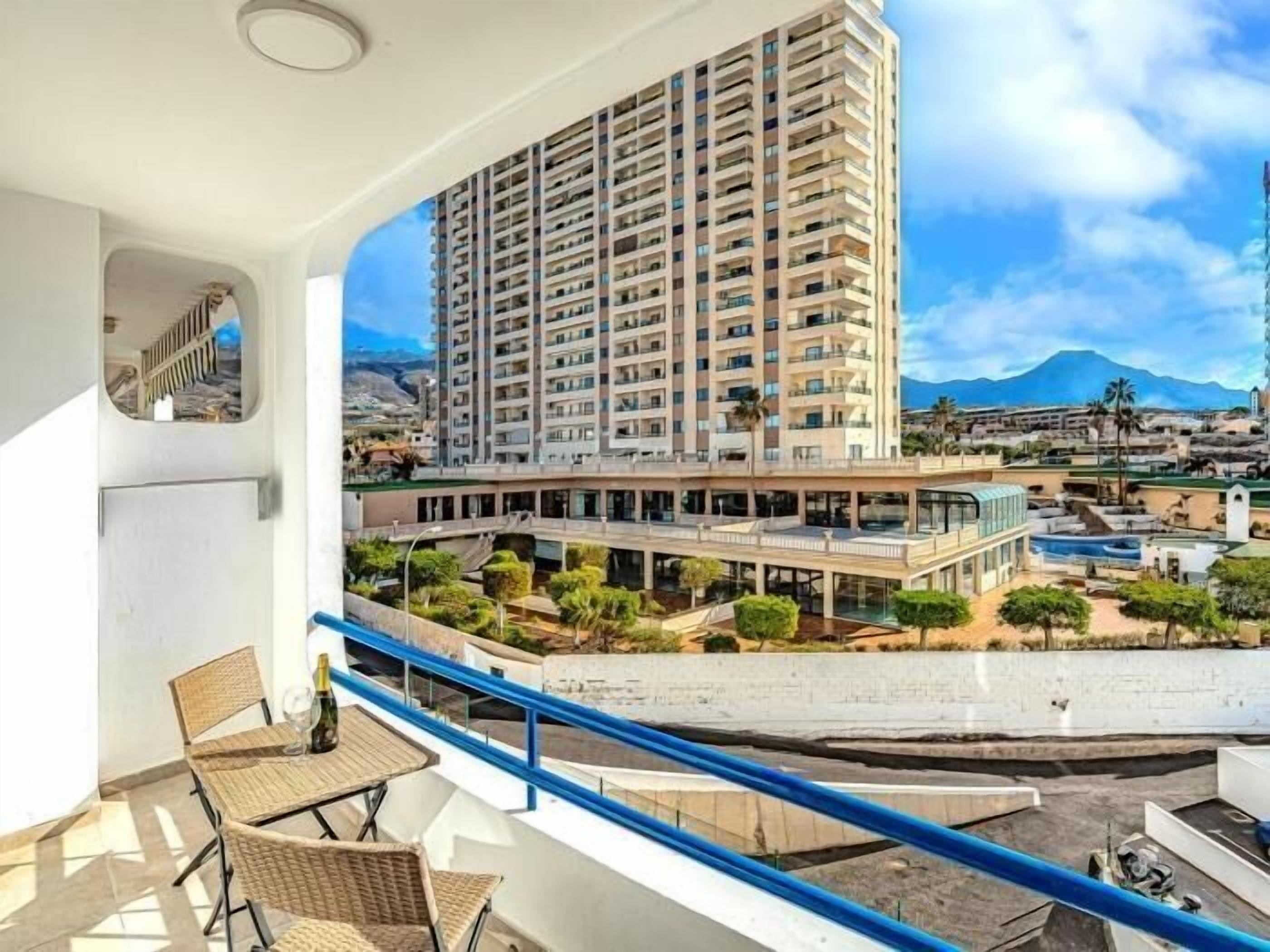 A balcony with wicker furniture and a bottle, overlooking a high-rise building and a pool area with greenery.