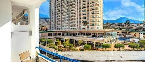 A balcony with wicker furniture and a bottle, overlooking a high-rise building and a pool area with greenery.