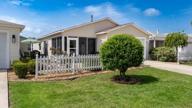 A single-story house with a white picket fence, a small tree in the front yard, and a clear blue sky.