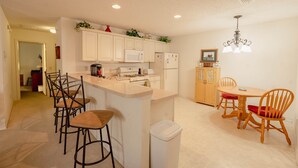 A kitchen with white cabinets, a microwave, a stove, and a trash bin. There are wooden chairs and a table with a chandelier.