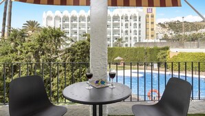Un balcón con dos sillas negras y una mesa, con vista a una piscina y un edificio blanco con balcones.