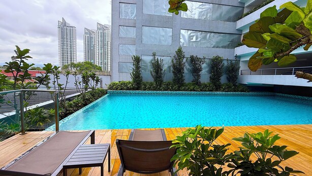Una piscina con lettini e un pontile in legno, circondata da vegetazione e moderni edifici sullo sfondo.
