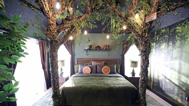 Ett sovrum med en säng placerad mitt under ett tak av konstgjorda träd, ett sängbord med en lampa och en vägg prydd med en väggmålning med naturmotiv.