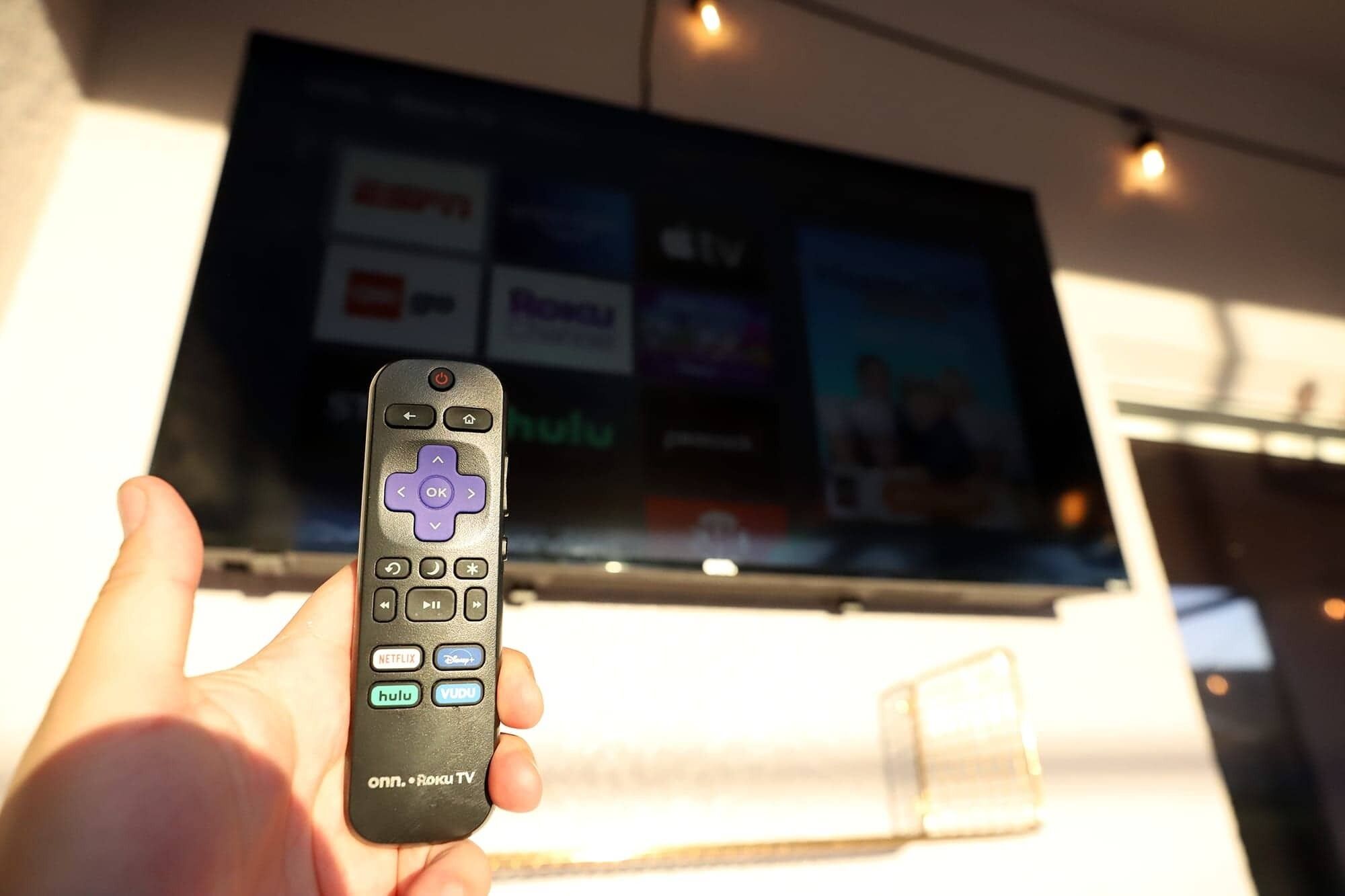 En hand håller i en fjärrkontroll, och i bakgrunden syns en tv-skärm som visar olika streamingtjänster.