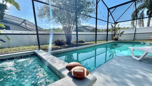 Una piscina con un balón de fútbol en el borde, rodeada por una zona techada con vistas a palmeras y un cielo despejado.