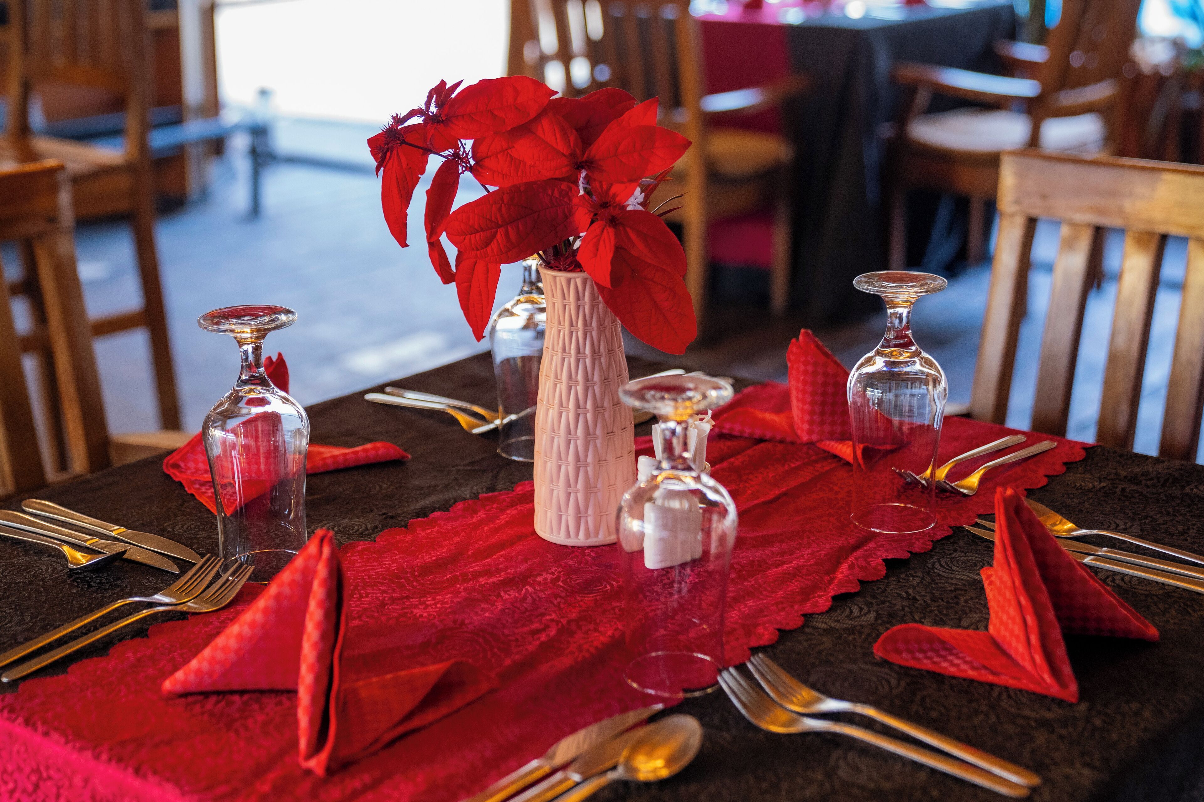 Une table à manger dressée avec des serviettes rouges, de la vaisselle transparente et un vase rempli de fleurs rouges.