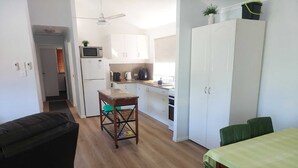 A compact kitchen with white cabinets, a microwave, and a refrigerator. There's a small table with a green chair, and a dining table with a green chair. - Palm Grove Cottage Su (Agnes Water)