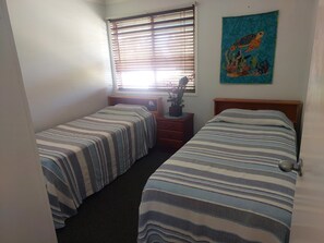 A room with two beds, a window with blinds, a nightstand with a lamp, and a wall painting.