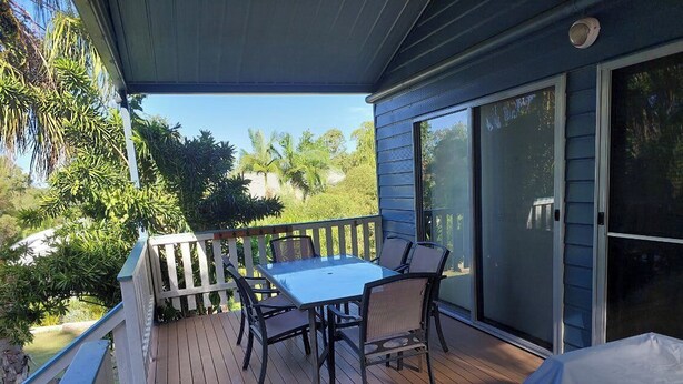 A covered outdoor area with a table and chairs, a sliding glass door, and a view of greenery. - Palm Grove Cottage Su (Agnes Water)