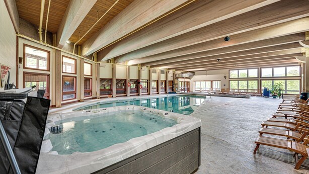 Una amplia piscina cubierta con un jacuzzi, vigas de madera en el techo y ventanas iluminadas por el sol.