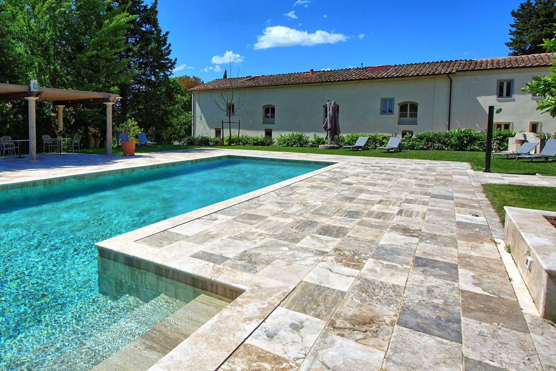 A swimming pool with a tiled deck, surrounded by a building with multiple windows and a pergola with chairs.