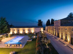 Une cour avec une piscine, des chaises longues et un bâtiment en pierre dont les fenêtres sont illuminées.