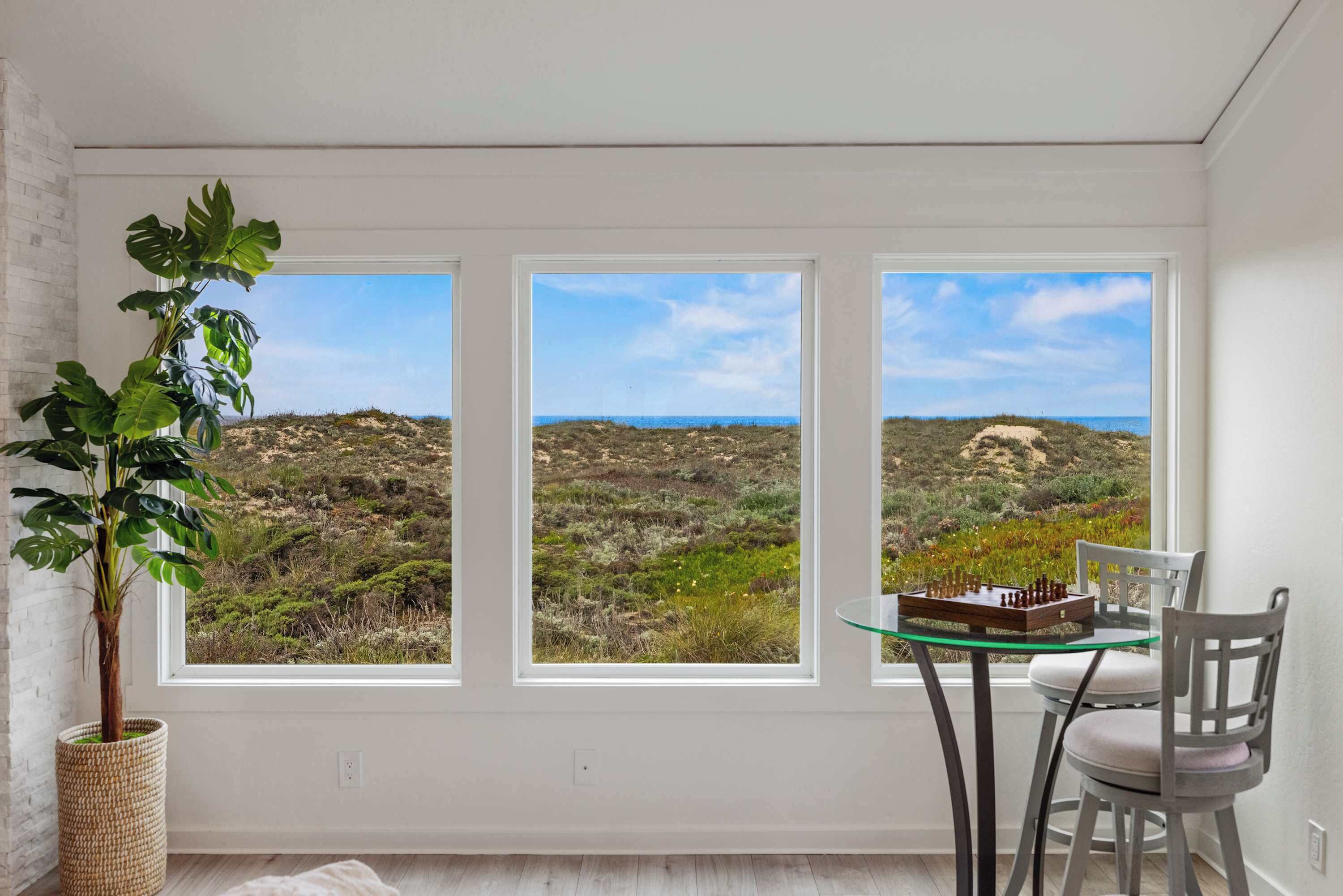 A room with three large windows, a potted plant, a glass table, and a chair.