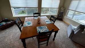 A dining area with a wooden table, four chairs, and two large windows with blinds. - Seaside Paradise at Sebastian Shores (Gold Beach)