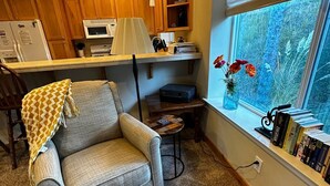 A cozy living room with a grey armchair, a small coffee table, and a bookshelf by a window with a view of greenery. - Seaside Paradise at Sebastian Shores (Gold Beach)