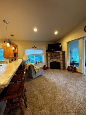 A spacious living room with a fireplace, a dining area with a long table and chairs, and a view of the outdoors through a window.