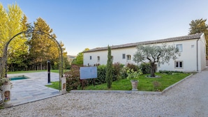 A white building with a tiled roof, a gravel pathway, and a small garden with trees and shrubs. - Borgo Ariano 18 – Apartment with pool (Volterra)