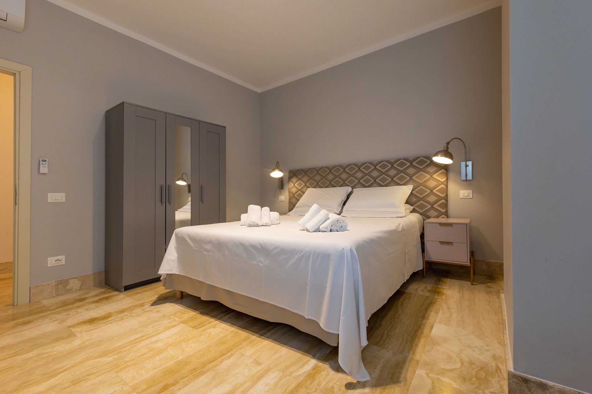 A hotel room with a bed, a wardrobe, a nightstand, and a wall-mounted lamp.