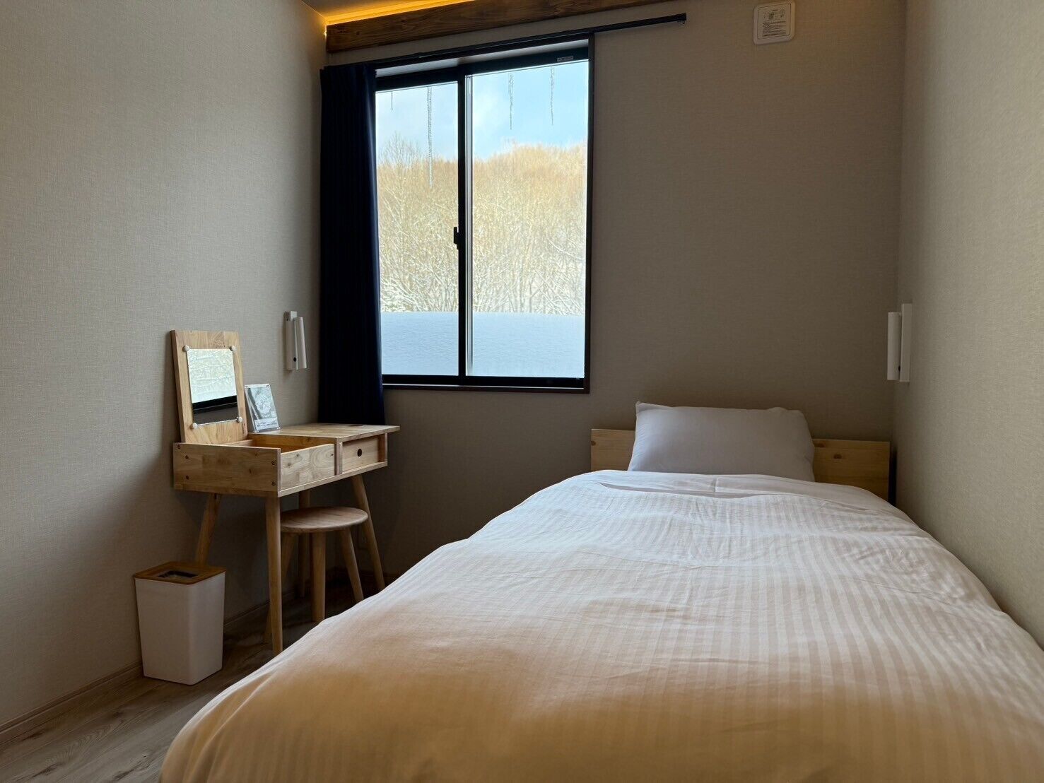 A bedroom with a bed, a desk with a chair, a window, and a small trash bin.