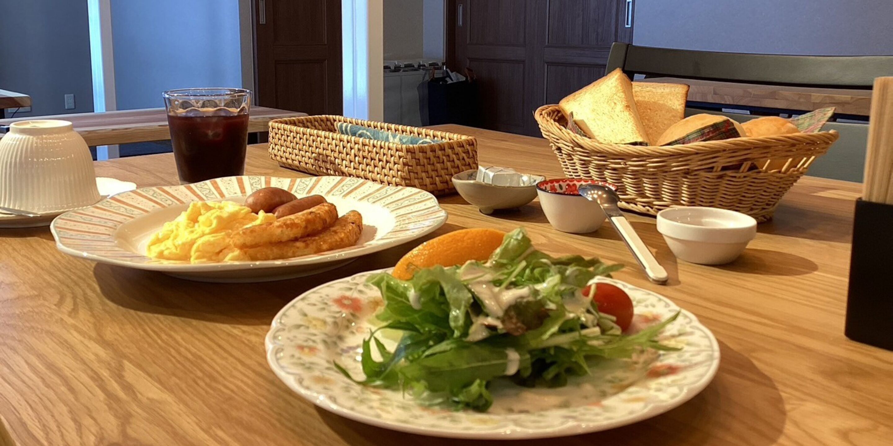 A breakfast table with plates of food, a glass of iced tea, and baskets of bread.
