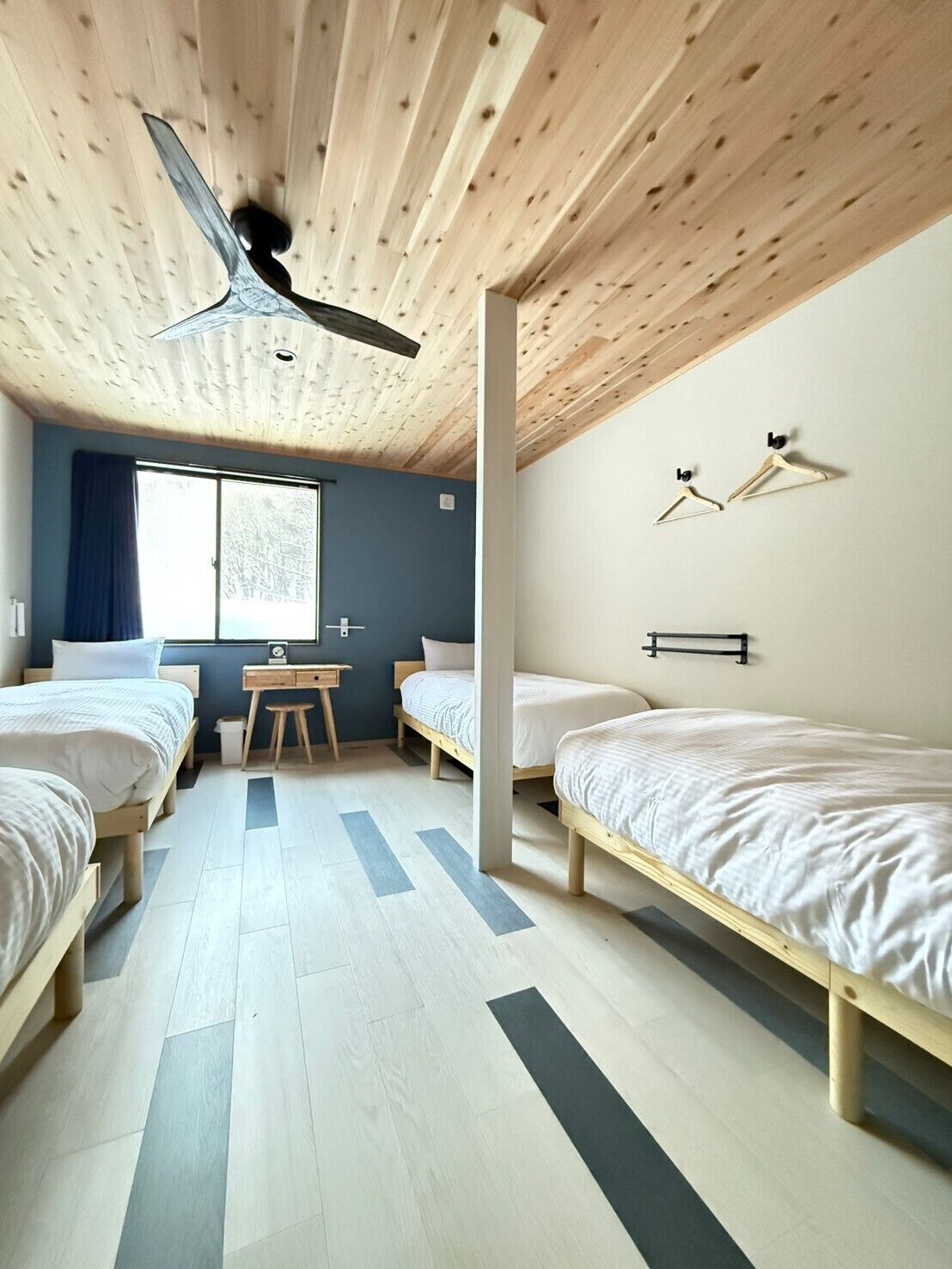 A modern bedroom with two beds, a wooden ceiling, a window with curtains, and a small desk.