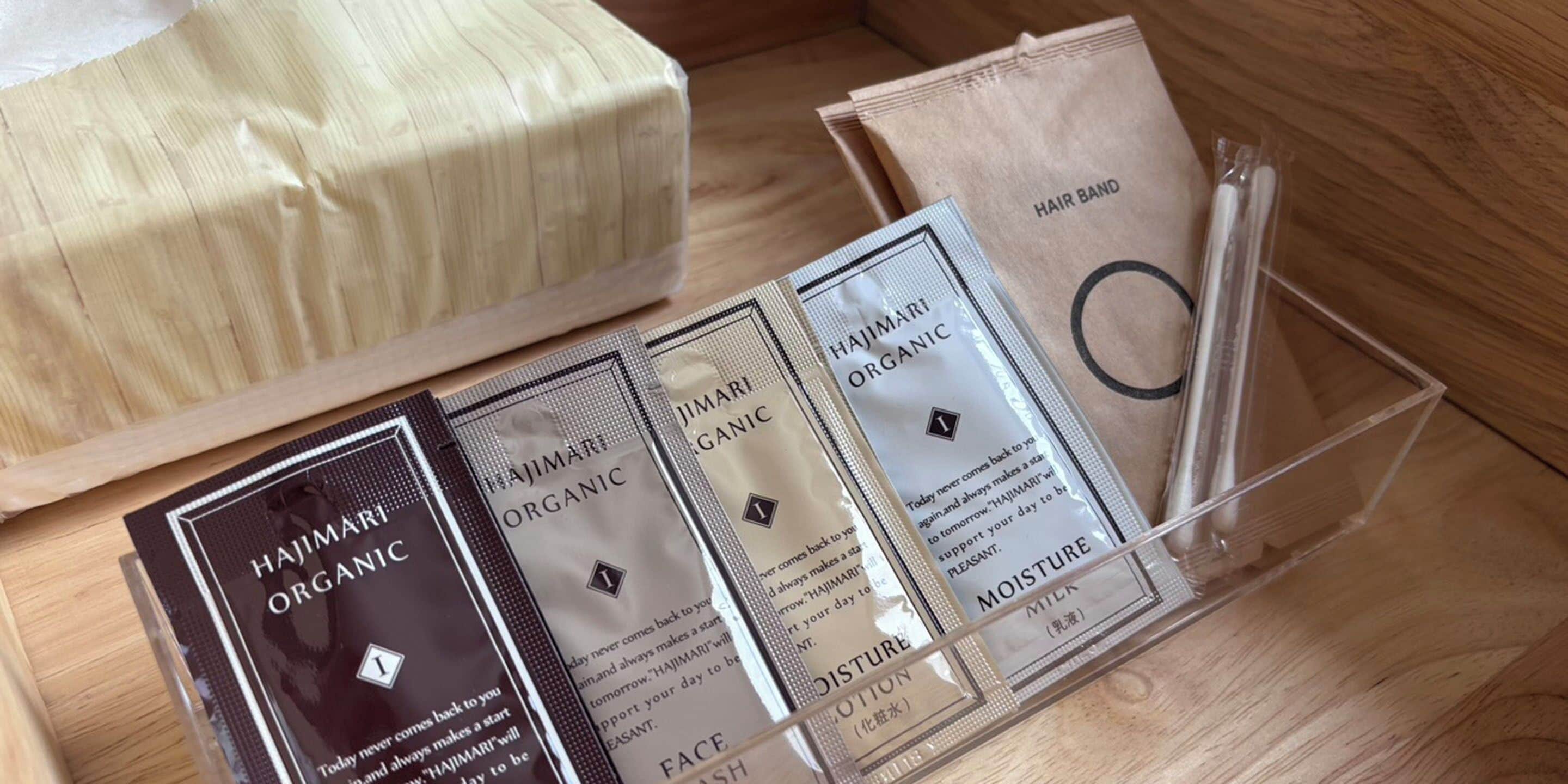 Superficie de madera con un organizador de plástico transparente que contiene varios objetos: una caja marrón con la etiqueta 'HAJIMARI ORGANIC', una bolsa de papel marrón con la etiqueta 'HAIR BAND' y un recipiente de plástico transparente con una etiqueta que dice 'MOISTURE (乳液)'.