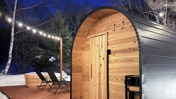Una cabaña de madera con techo curvo, una puerta de madera y dos sillas plegables en una terraza de madera.