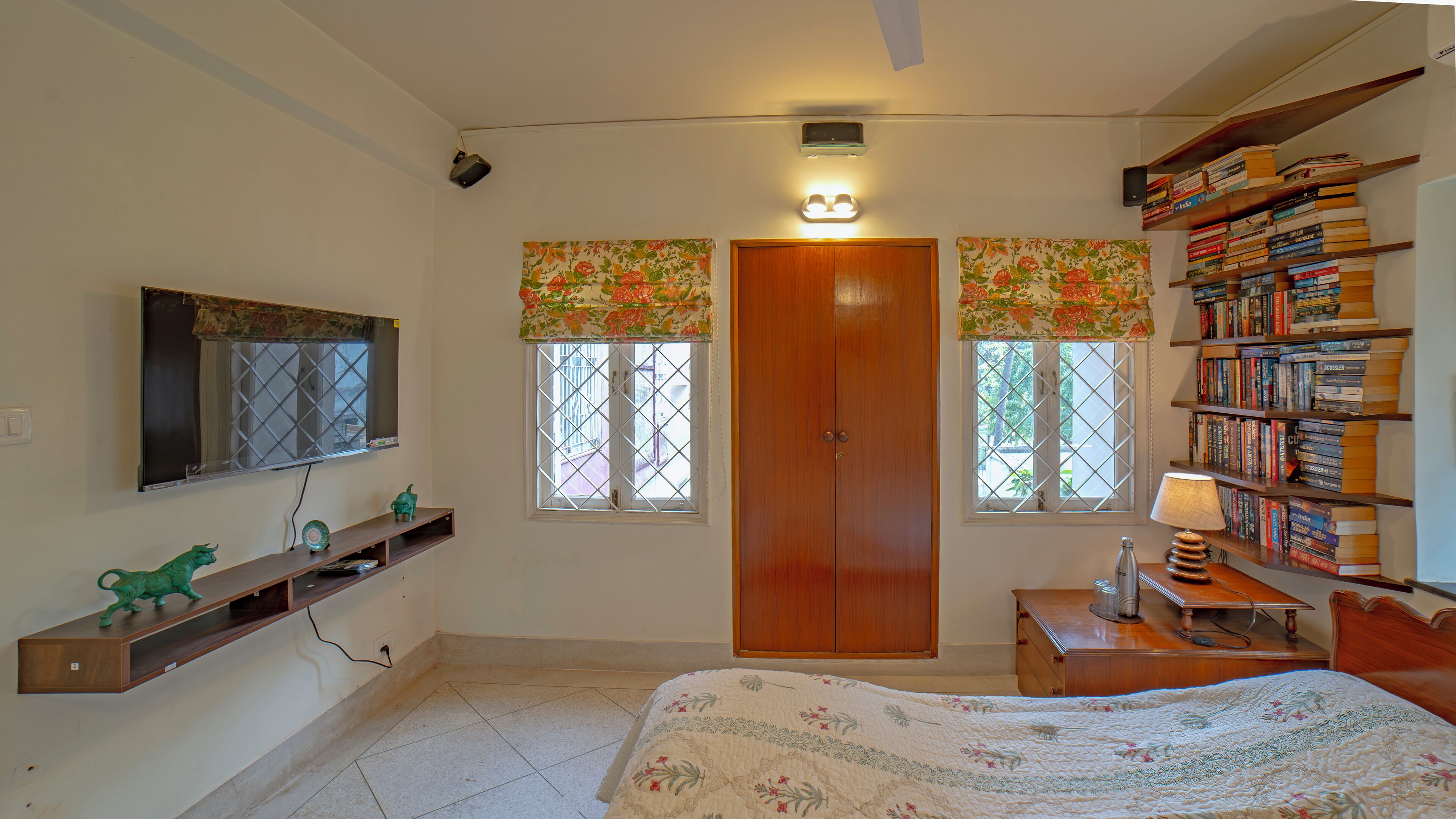 Un dormitorio con cama, un armario de madera, una estantería con libros, un escritorio con lámpara y dos ventanas con cortinas florales.