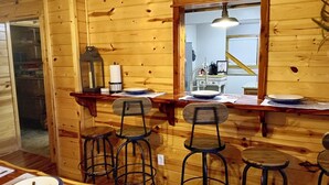 Interior de una cabaña de madera con comedor, que incluye una mesa de madera, cuatro taburetes altos y un espejo sobre la mesa.