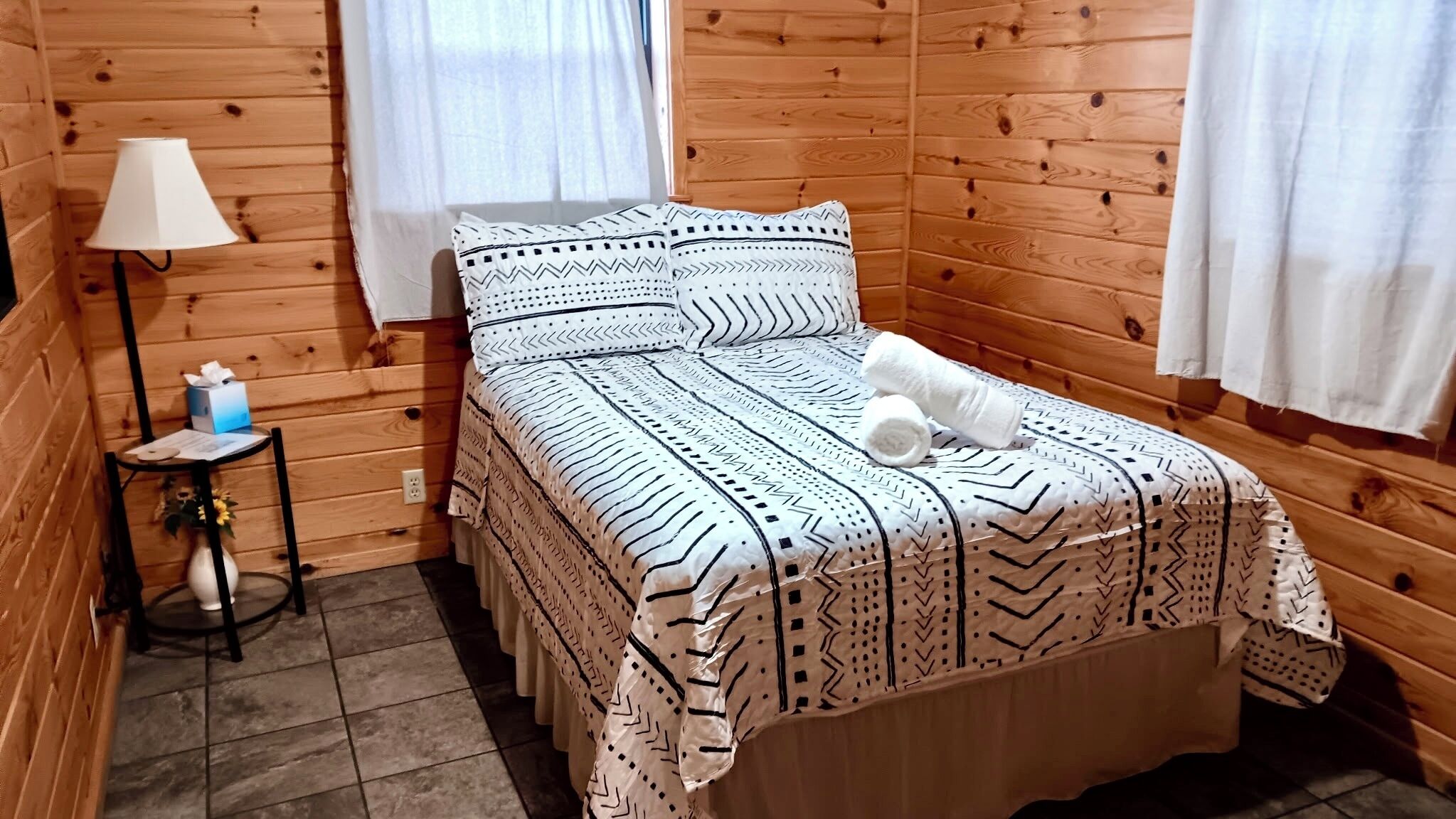 A wooden cabin room with a bed, a nightstand, a lamp, and a window.