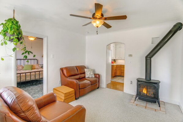 A living room with a leather sofa, a brown armchair, a hanging plant, a ceiling fan, and a wood-burning stove. - Farmhouse w/ Horse Corral Near Wind River Arena (Rigby)