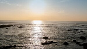 Ein Strand mit Sonnenuntergang über dem Wasser, Felsen im Vordergrund und einem klaren Himmel.