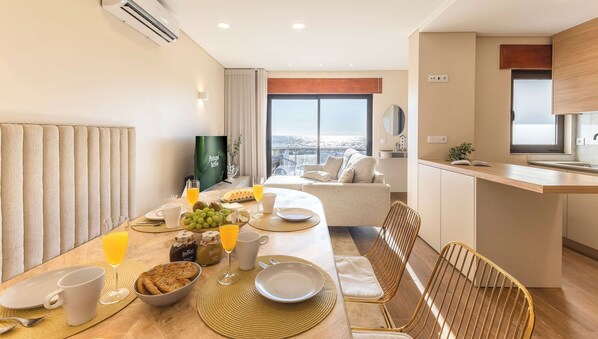 A hotel room with a dining table set for breakfast, a sofa, a kitchen area, and a large window with a city view.