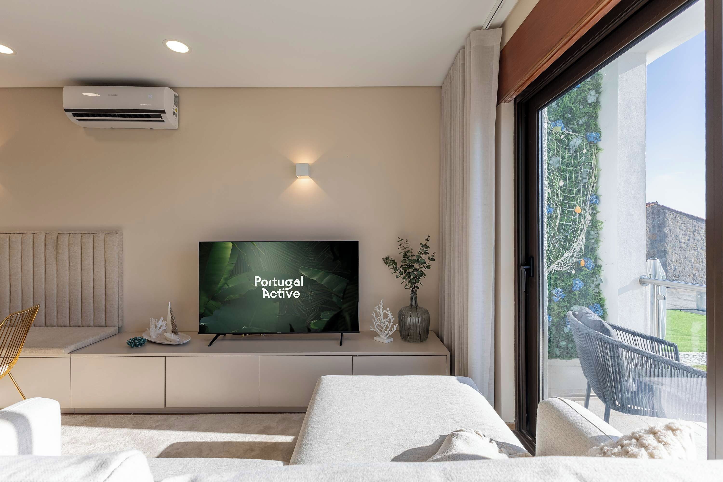 Un dormitorio moderno con televisor, una mesita de noche con objetos decorativos, una ventana con vistas a zonas verdes y un edificio, y aire acondicionado instalado en el techo.