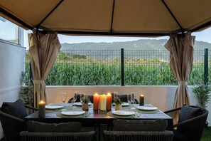 An outdoor dining area with a table set for two, surrounded by wicker chairs, under a canopy.
