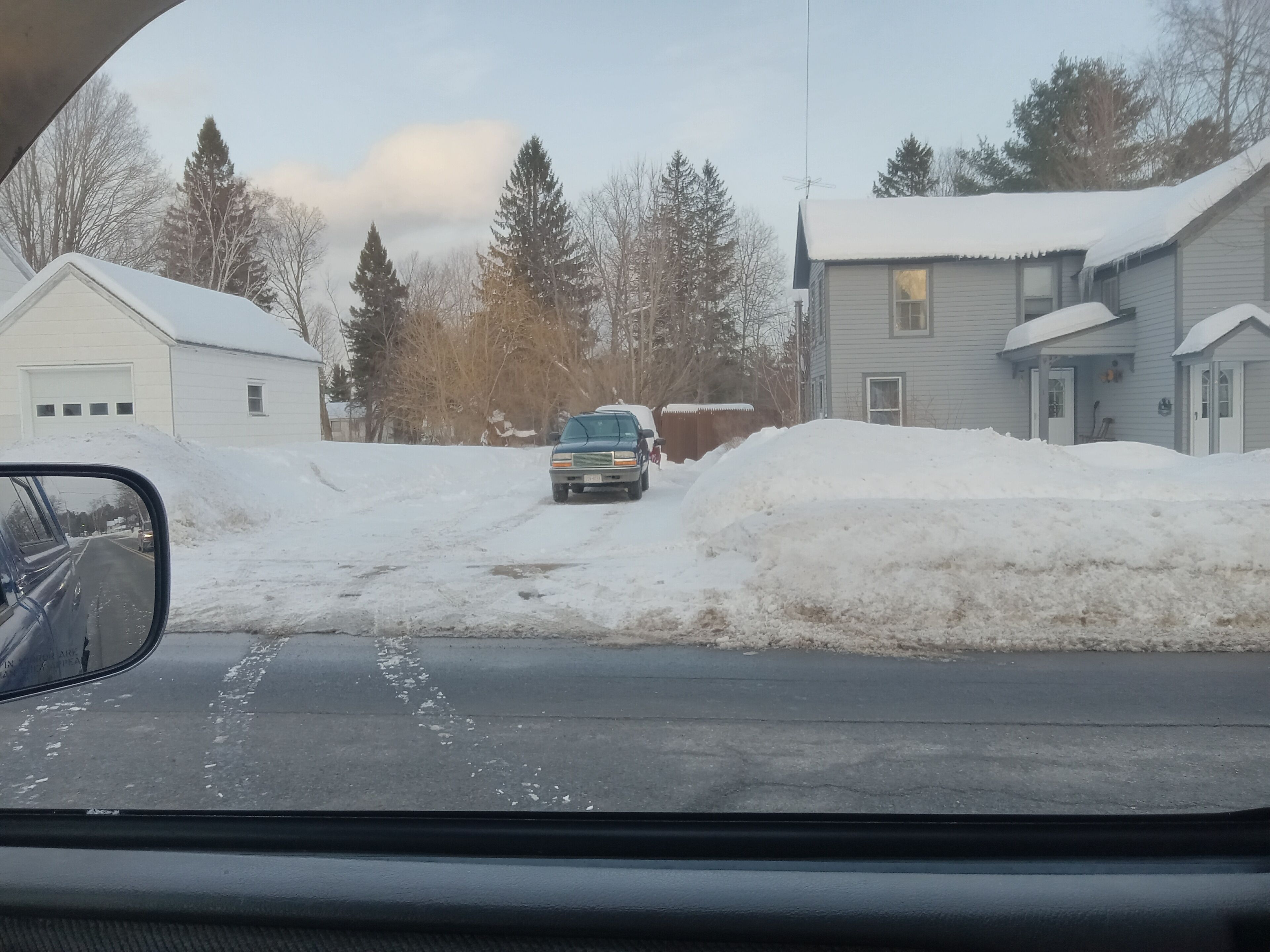 A residential street with houses on both sides, a parked truck, and a snow-covered road.