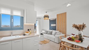 Eine moderne Küche mit großem Fenster, einem Essbereich mit weißem Tisch und Stühlen sowie einem Wohnbereich mit Sofa und dekorativer Wandverkleidung.