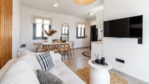 Ein modernes Wohnzimmer mit Sofa, Essbereich mit Tisch und Stühlen, Küche mit an der Wand montiertem Fernseher und einem großen Fenster, das für natürliches Licht sorgt.