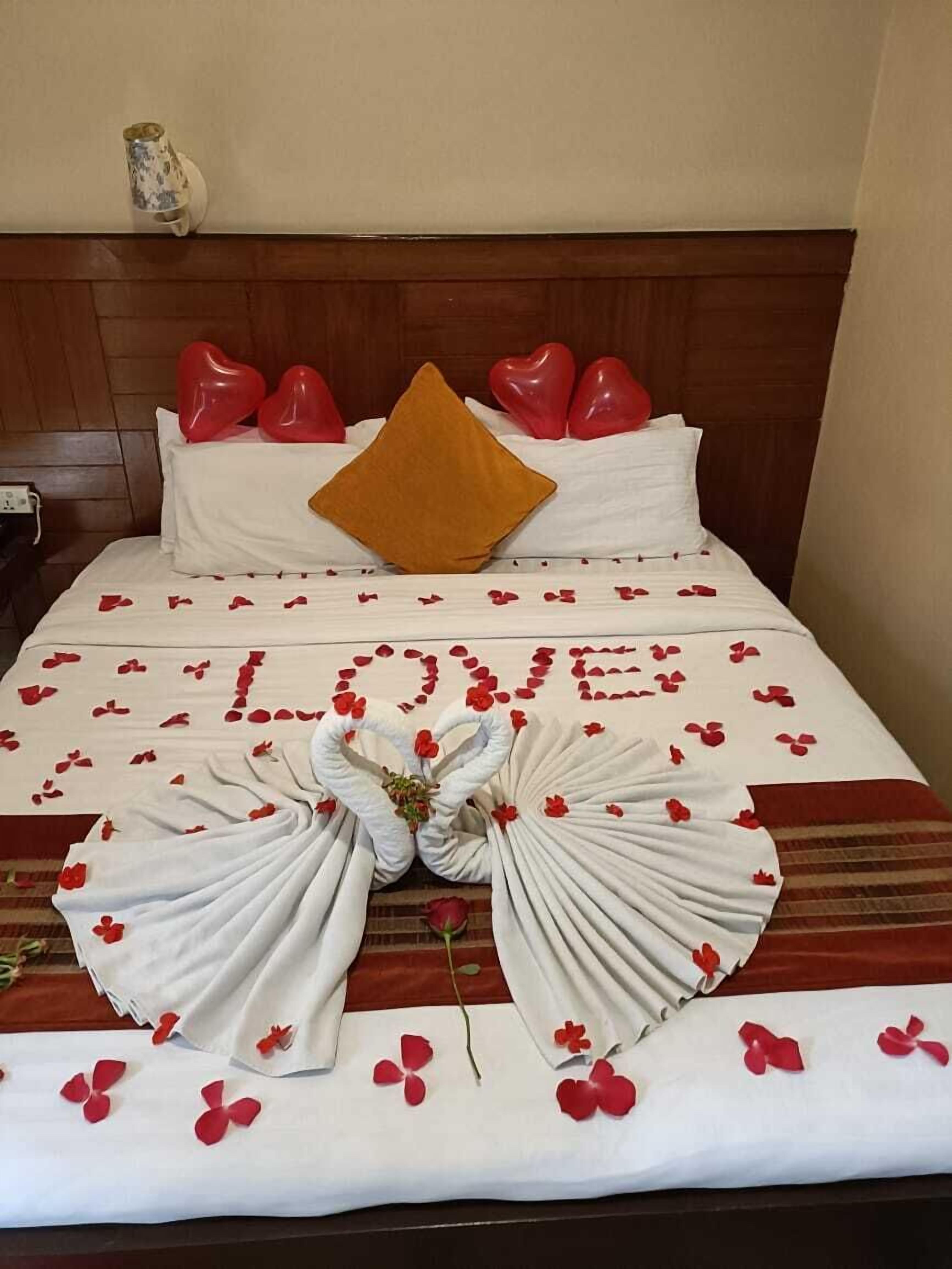 A bed with a heart-shaped pillow arrangement, a large 'LOVE' decoration made of rose petals, and a bouquet of flowers.