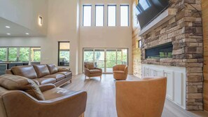 A spacious living room with a stone fireplace, leather sofas, and large windows offering a view of the outdoors. - Luxe Lakefront Retreat w/ Hot Tub & Game Room (Rogers)
