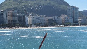 En vaffel med chokolade på en tallerken, et glas kaffe og udsigt til en strand med bygninger og et bjerg i baggrunden.
