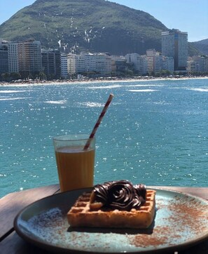 Gofre con chocolate en un plato, una taza de café y una vista a la playa con edificios y una montaña al fondo.