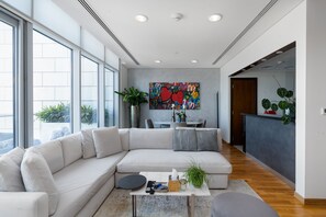 Ein modernes Wohnzimmer mit einem großen, weißen Ecksofa, einem Couchtisch mit Fernbedienungen und einem farbenfrohen abstrakten Gemälde an der Wand.