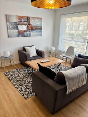 Ein modernes Wohnzimmer mit einer Couch, einem Couchtisch und einem Sessel. An der Wand hängt ein Gemälde und das Fenster bietet Blick auf Gebäude.