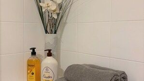 A bathroom with a white tiled wall, a white vase with white flowers, two bottles of toiletries, and a stack of grey towels. - Pinvidik apartment with private parking close to city center and train station (Saint-Brieuc)
